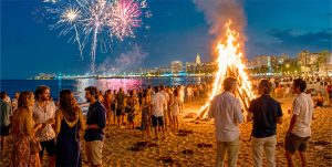 Hoguera en la playa durante la noche de San Juan 2026, con fuegos artificiales y ambiente festivo.